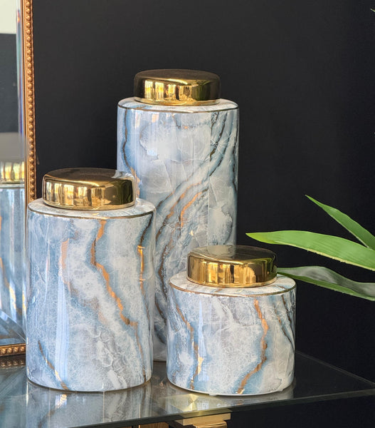 Trio of blue marble ceramic canisters with gold lids, styled on a glass table with a black background and decorative accents.