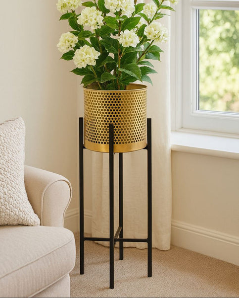Floral arrangement in a metal planter on a black metal stand next to a beige armchair.