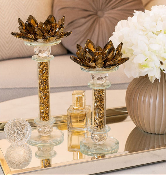 Decorative candle holders on a mirror surface with flowers and perfume bottle in the background