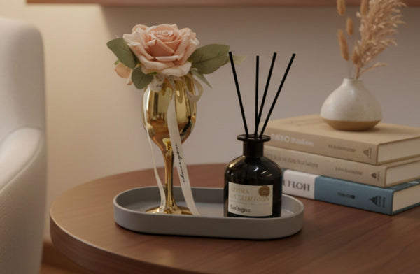 Decorative items including a gold champagne glass with a pink rose, a black diffuser bottle on a grey tray and books on a wooden table.