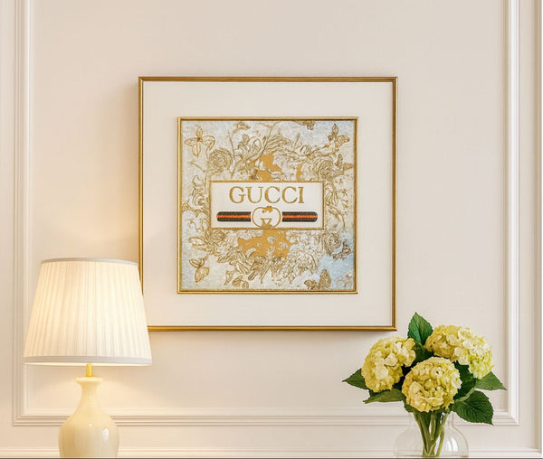 Framed Gucci artwork on a wall with a lamp and flowers in the foreground.