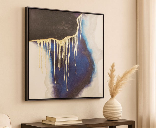 Abstract art piece with gold and blue elements on a beige wall, accompanied by a vase and books.