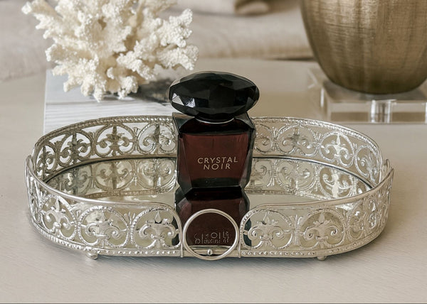 Decorative silver oval tray with a black perfume bottle labeled 'Crystal Noir' on a light surface.