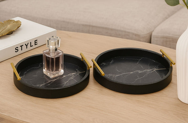 Two black marble textured trays with gold handles on a wooden surface, one holding a perfume bottle.