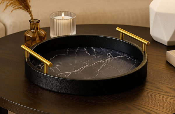Black marble textured tray with gold handles on a wooden table, surrounded by decorative items.