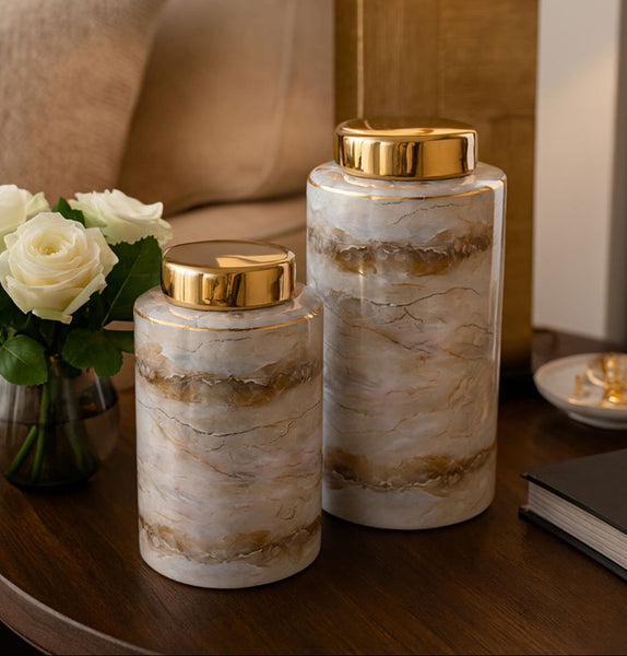 Two marble-patterned jars with gold lids on a wooden surface, with flowers and a bowl in the background.