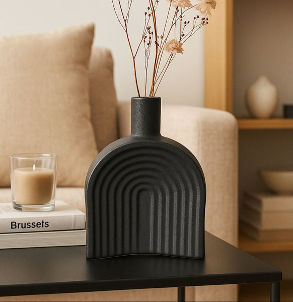 Black ceramic vase with arches holding small light pink flowers on a wooden surface.