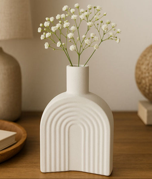 White ceramic vase with arches holding small white flowers on a wooden surface.