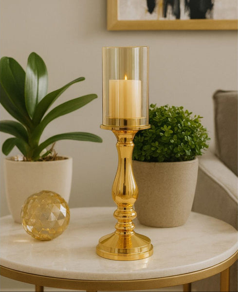 Gold candle holder with a lit candle on a marble table with plants and a gold decorative object.