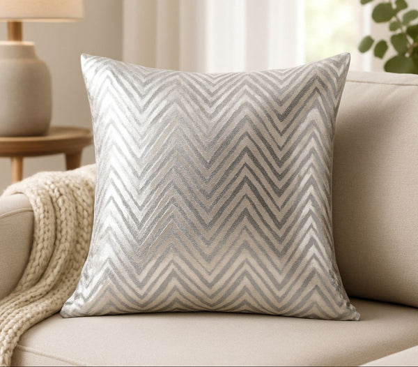 Gray and Silver chevron patterned pillow on a beige couch with a side table and lamp in the background.