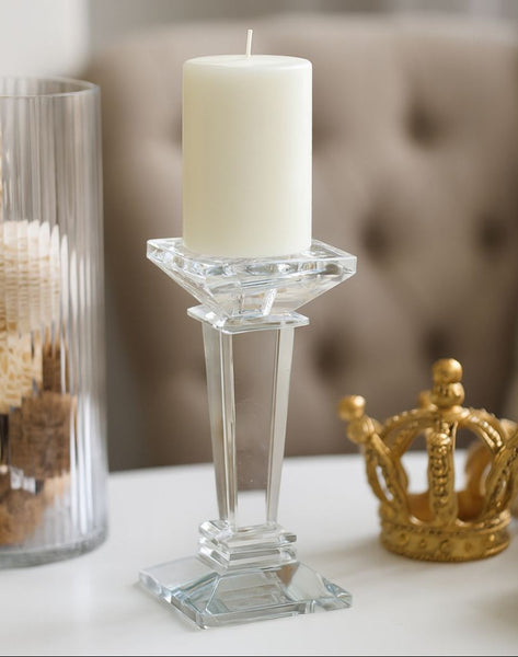 Clear glass candle holder with a white candle on a surface, with a decorative crown and jar in the background.
