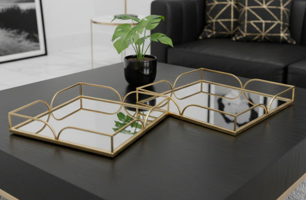 Two gold square mirror decorative trays on a dark surface with a plant and couch in the background.