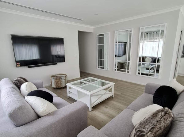 Hampton’s style white timber and glass coffee table styled between two grey fabric sofas.