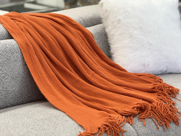 Cozy Levi Throw Rug Blanket in orange draped over a grey sofa with white cushion, showcasing soft texture and fringe.
