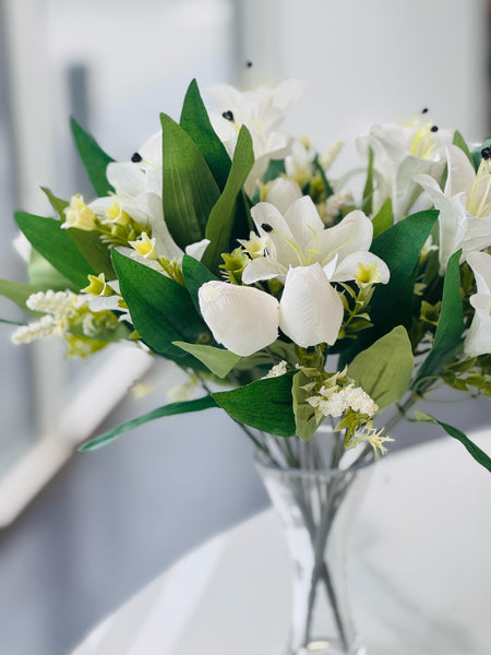 Close-up of white lily and tulip bunch with lush green leaves in a glass vase.