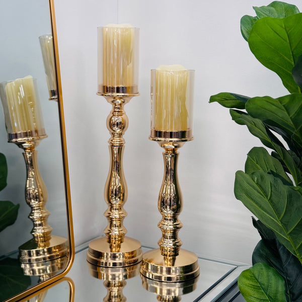 Gold pillar candle holders styled on a mirrored surface with lush green plants and reflective background, creating a luxurious home décor setting