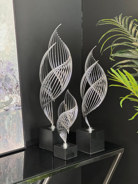 Elegant silver Ribbon Table Décor set in small, medium, and large sizes displayed on a black console against a dark backdrop with greenery – showcasing a modern and artistic interior accent.